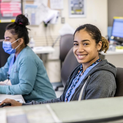 Image of A healthcare worker with her hair tied back in a ponytail sits at her desk and smiles for the photo