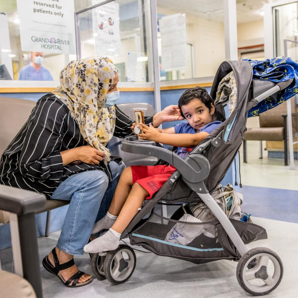 A child sit in a stroller while waiting for care at that Hospital. Their parent is leaning in to stroke their hair. 